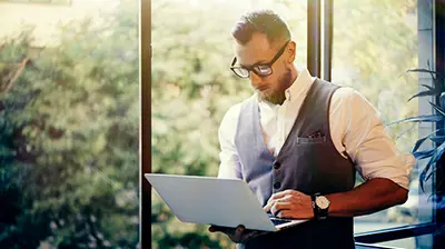 Man dressed in business casual attire standing by window while using a computer.