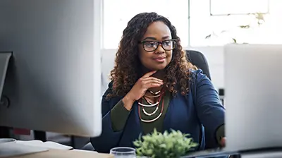 A woman sitting in front of two computer screens, looking at one screen with a hand under her chin 