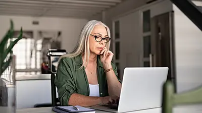 A woman sitting at her kitchen table and using a laptop while resting her hand on her chin 