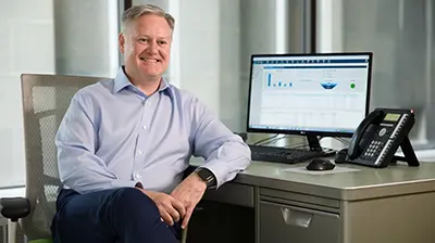 Man dressed in casual attire sitting at desk looking forward and smiling.