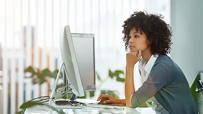 Woman in casual attire sitting at a desk in an office, she is looking at a large computer screen in front of her. 